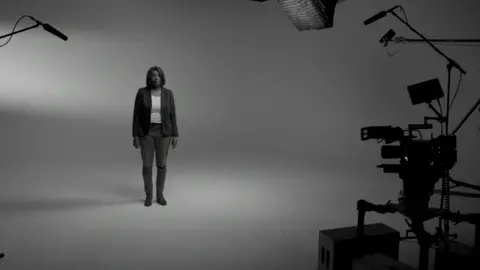'Change Takes All' - Cocoa Media TV An image from the documentary Change Takes All. Jennifer Donaldson is pictured in black and white. She can be seen standing in a studio on her own. In front of her there are cameras, lights and sound equipment. She is wearing a black jacket, jeans and a white top. Her hair is dark and cut in a bob.