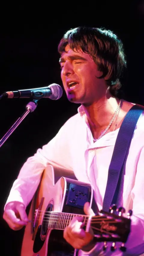 Getty Images Noel Gallagher playing a guitar and signing on stage next to a microphone in a stand. He is wearing a white long-sleeved top.