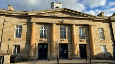 Durham Crown Court. A two storey Georgian court building made form yellow stone with four columns at the front, a small clock tower and long rectangular windows
