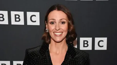 Getty Images A woman in her fourties with brown hair in a curled and styles bob. She wears a black shirt or blazer with tiny rhinestones spread across it. Behind her is a black board with the BBC logo on it in white.