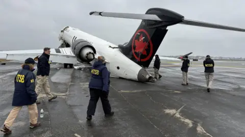National Transportation Safety Board (NTSB) investigators are seen on the tarmac walking across behind the back of the plane. It is stormy outside inspect the wreckage of an Air Canada Express jet that collided with a fire truck at LaGuardia Airport in Queens, New York, U.S., March 23.