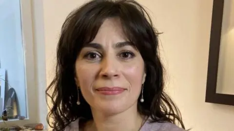 A woman with dark hair and earrings in a room with a picture on the wall to the side of her.