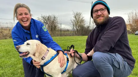 RSPCA Kim Walters and Andy Cook, with Daisy. Both are smiling at the camera while crouched down on some grass and holding the dog. Kim has blonde hair and is wearing a bright blue RSPCA jacket. Andy has glasses and a beard and is wearing a winter hat and dark fleece.