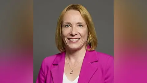 UK Parliament Helen Morgan has a blonde bob and is smiling. She wears a white top and bright pink suit jacket. She has a silver necklace on and is posing in front of a grey background.