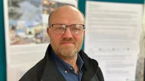 A man with a beard and glasses wearing a denim shirt under a dark jacket stands in front of information boards inside a village hall