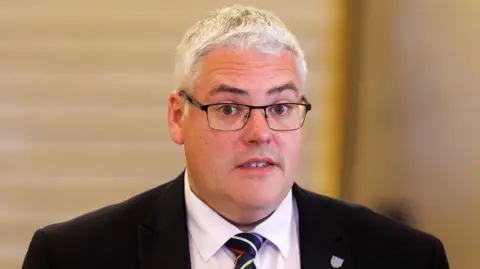 PA Media Gavin Robinson, dark suit, stripy tie. He has glasses and grey hair
