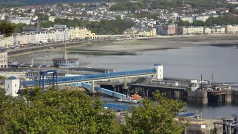 A view of Douglas Bay on a summer day, affording a sweeping view of terraced buildings, and a sandy, seaweed-laden beach at low tide. At the forefront of the image stands a long blue windowed walkway, slipways and a pier forming the Sea Terminal port.