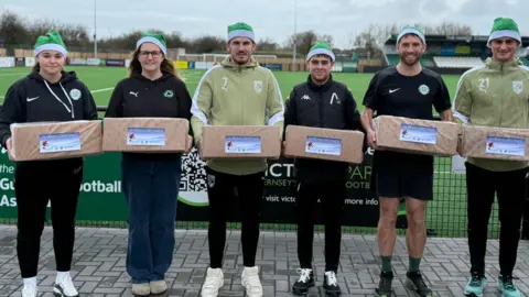 Victoria Park Two women and four men dressed in sports kit are standing on front of a football pitch. They are all wearing green santa hats and holding boxes wrapped in Christmas paper, with greetings tags on them.