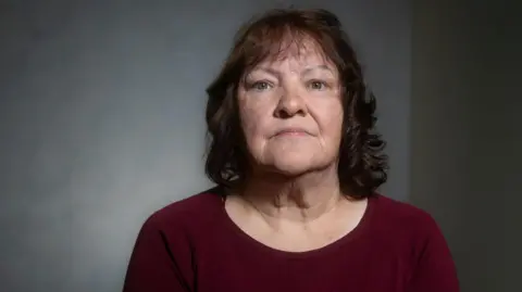 A woman sitting against a softly lit, neutral background, looking toward the camera with a calm, serious expression and wearing a burgundy top.