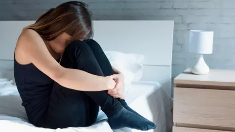 Getty Images A stock image of a woman with long dark hair with slightly lighter streaks, sitting on a bed with her knees pulled up to her head and her arms clasped around her legs.