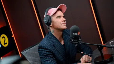 A man, Robbie Williams, in the BBC Radio 2 studio wearing a pink cap and pin striped suit.