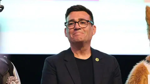 Getty Images Burnham smiling, reacting to his victory in the Makerfield by-election. He is wearing a black top, black blazer and black framed glasses. He has dark brown hair.