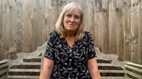 Zoe Dennis/BBC Nicki Duffield sitting on a wooden bench looking at the camera. Behind her is a wooden fence. She is wearing a black and white leaf-pattern dress. She has blonde hair and glasses.