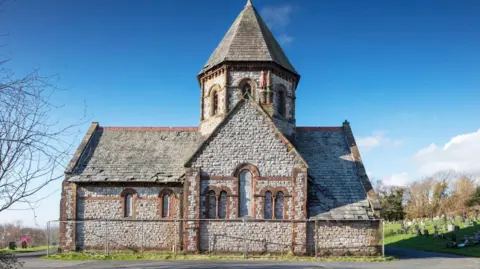 A chapel in Barrow Cemetery on a sunny day. The building has windows but they have been boarded up. The building is Victorian-style with large white and red bricks. 