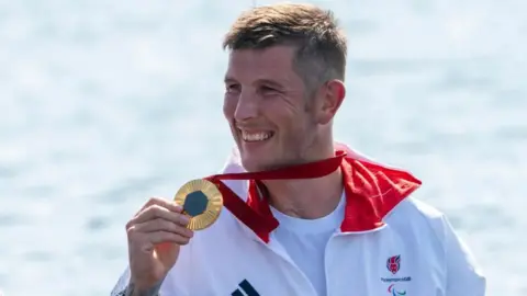 PA Media Paralympian Gregg Stevenson holding up a gold medal and smiling in the sunshine. He is wearing a white tracksuit and is standing in front of a large body of water
