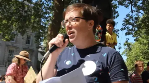 Clare Clare from Berkshire is holding a microphone as she speaks to a crowd. She is wearing a blue T-shirt and has short, brown hair and is wearing tortoiseshell glasses.