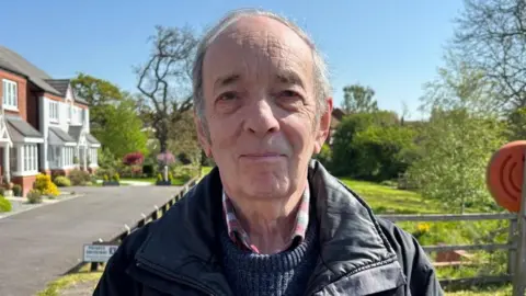 There is a man who has short grey hair, a black coat, dark blue jumper and a red, white and grey checkered shirt. He is stood on a pavement, in front of a private driveway and a park, which includes green trees, grass and yellow wildflowers.
