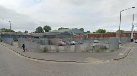 Google An industrial area with fenced parking, warehouse‑style buildings and a person walking a small dog along the pavement at a street corner, with trees visible behind the buildings.