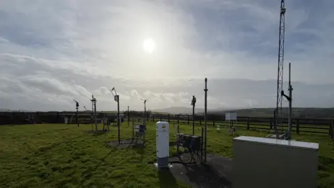 Bruce Griffith The picture shows a weather monitoring site set up in an open grassy field. There are multiple scientific instruments on metal poles, small towers and enclosed units, all designed to measure different aspects of the weather. Some are tall masts with sensors fixed to the top, while others are shorter devices closer to the ground. The area is fenced off, and the field stretches towards distant hills under a bright, hazy sun. 
