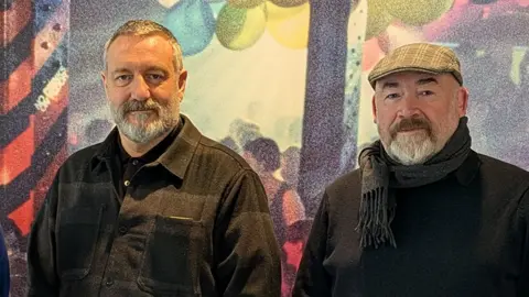 Martin Green and James Lawler in the exhibition space. They are both wearing dark jackets and both have short grey beards