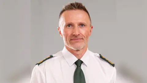 Welsh Ambulance Service Jason Killens looking at the camera smiling. He has short cropped hair and is wearing a white shirt with a green tie and green and gold embossed ambulance shoulder badges.