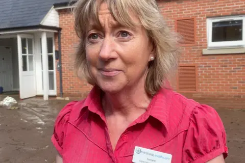 Helen Bayliss has blonde hair and is wearing a pink top, she is standing in front of a building and low level flood water is visible near the ground