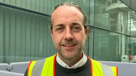 Matt Knight/BBC A head and shoulders picture of Colin Walsh wearing a high-vis jacket inside a building at Stansted Airport. His hair is swept back and he has facial stubble. He is wearing a white shirt under a grey crew-neck jumper.