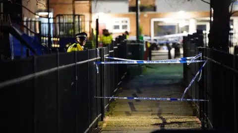 PA Media A night shot of the police cordon and police officers standing on one side of a black gate.