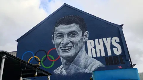 Mural of Rhys McClenaghan. It has a blue background and the Olympic rings on the left side with 'RHYS MBE' on the right. 