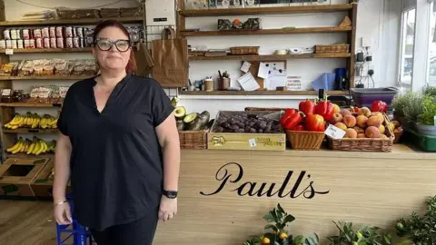 Clare Dutton / BBC A woman in a black top stood in a fruit and vegetable shop with fruits and vegetables on display.