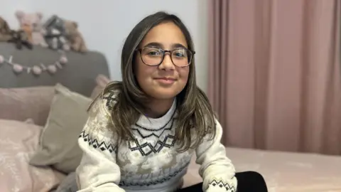 BBC/Kate Bradbrook A young girl with brown hair and glasses sits on her bed, smiling at the camera.