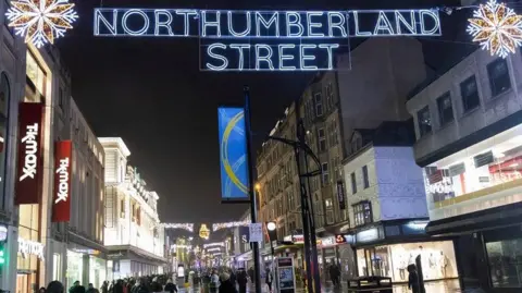 Newcastle City Council Bright Christmas lights are illuminated over the busy pedestrianised street. Shops line either side of the walkway. One display overhead shows the words: "Northumberland Street".