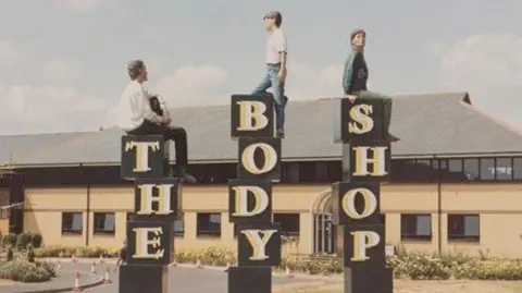 Giles Penny Three human sculptures are positioned outdoors on tall black blocks and arranged to spell “THE BODY SHOP.” Each figure sits on top of one of the blocks. The sculptures are placed in front of a large building with a sloped roof and arched entrance. The scene includes a lawn, a driveway with traffic cones, and a partly cloudy sky.