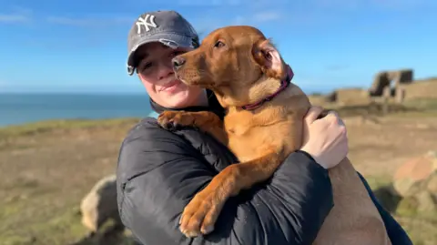 A woman standing at Grosnez holding a puppy with the landscape and sea behind her.