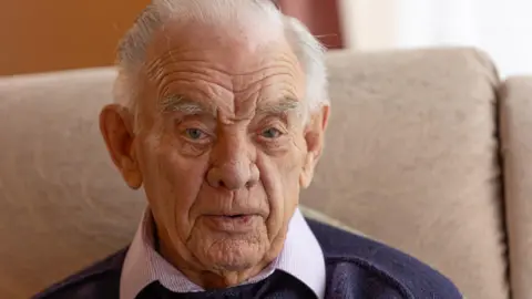 A close up of Barry. He has short grey hair and is wearing a navy jumper, with a pink shirt underneath.