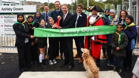 GWR A group of people, including West of England mayor Dan Norris and local MP Darren Jones, stand in front of a green ribbon at the opening of the new Ashley Down Station in Bristol. Local schoolchildren are also pictured, as is the city's lord mayor and Dan Norris's dog