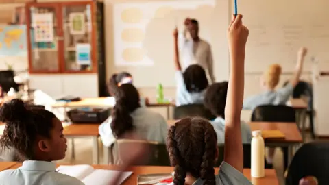 A classroom with children putting their hands up to answer a question posed by a teacher.