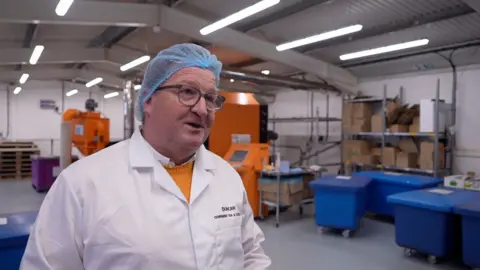 A man in a white coat smiles, behind him are large machines for coffee roasting and large blue bins that contain the beans.
