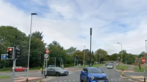 The junction of Eastfield Road with Eye Road in Peterborough. It shows at least seven traffic lights and cars manoeuvering through it. There are wide grass verges and trees on the left. 