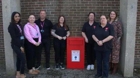 Several people look at the camera next to a brick wall, which in the middle of them has a red metal container attached to it. It has door or drawer at the top that knives can be put in to