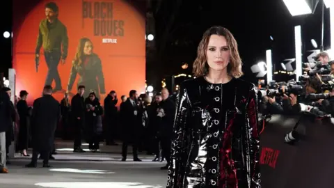 PA Media Keira Knightley successful  an all-black outfit with photographers and a ample  promotional representation  connected  a partition  astatine  the premiere of Black Doves successful  London connected  3 December