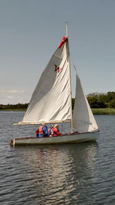 A small white sailboat with two large sails is in the middle of a calm lake. Three people wearing red life jackets sit in the boat, whilst trees line the background. 