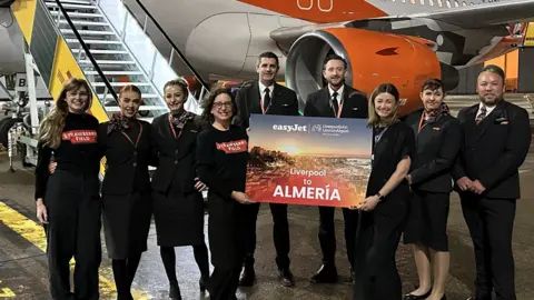 Liverpool John Lennon Airport A group of nine men and women stand in front of a plane with orange Easyjet signage on it. Most of them are wearing formal suits, while two of the women wear black t-shirts with the words 'Strawberry Field' on them. Two women at the front hold a sign which says 'Liverpool to Almeria'. 