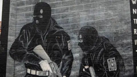 A black and white mural painted on a wall in east Belfast. There are two men, dressed in black, with black balaclavas and holding guns.