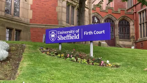 A purple sign says "University of Sheffield, Firth Court". It is on grass outside a red brick building with arched windows.