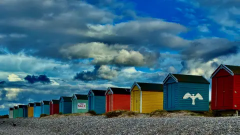 Gavin Neil Brightly-coloured beach huts - red, blue, yellow and green - under a cloudy blue sky.