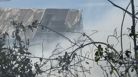 meal roof of a whare house is falling down as plumes of grey smoke engulf the building