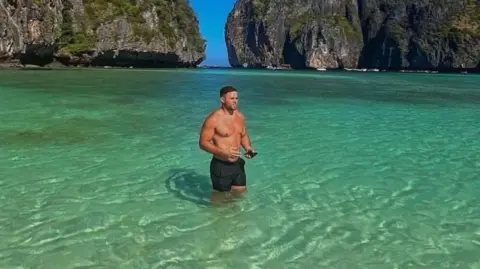 Family photo Corey Beavis pictured in black swim shorts standing in clear blue green water with mountains behind