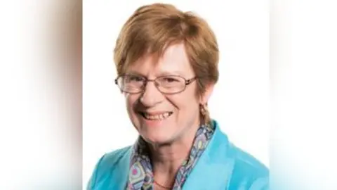 Senedd Cymru A woman with short brown/red hair, wearing glasses and a light blue jacket, smiles at the camera 