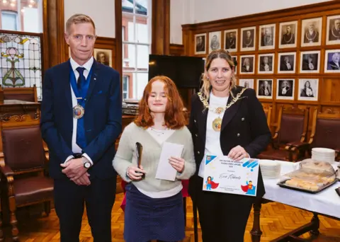 Evie, a teenage girl with short red hair, holds an award as she stands in the Douglas council chamber between Mayor Natalie Byron-Teare and consort Andy Teare.